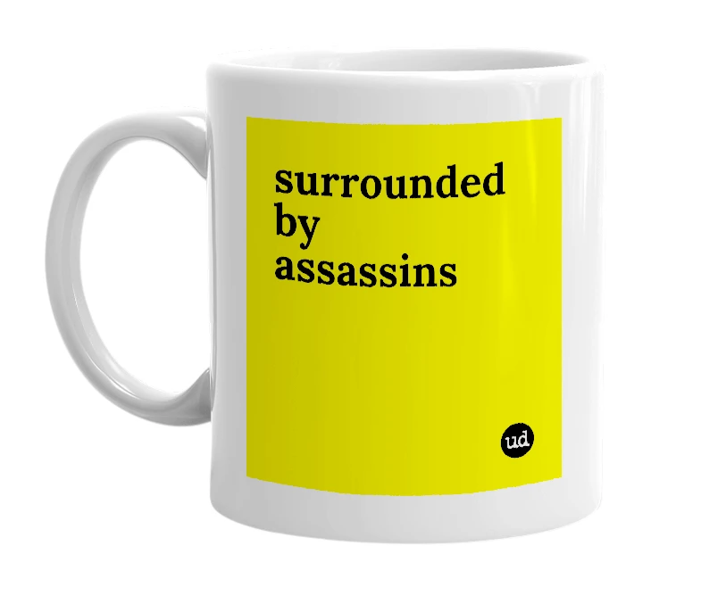 White mug with 'surrounded by assassins' in bold black letters